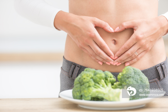 Woman with sports figure on her belly shows heart shape. Fresh broccoli in plate on kitchen table