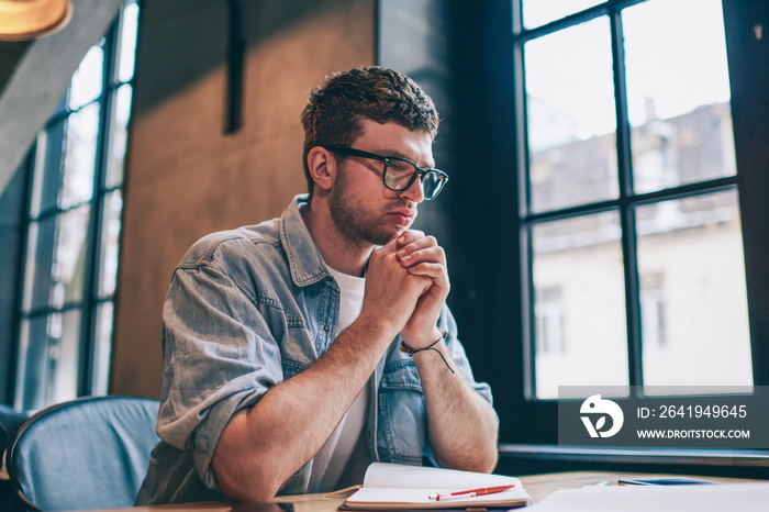 Puzzled male author in spectacles for vision correction thinking on ideas for article sitting at des