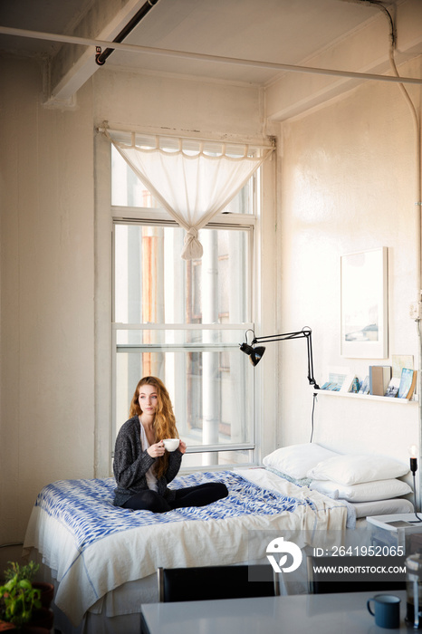 Woman with cup looking away while sitting on bed at home