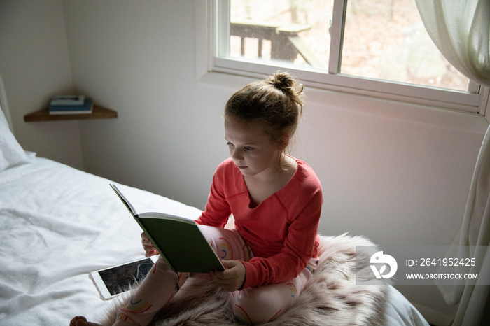 High angle view of serious girl reading book while sitting on bed against window at home