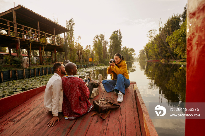 Smiling young woman taking pictures of men with digital camera in trajinera boat