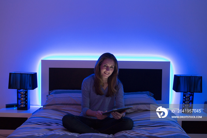 Portrait smiling woman using digital tablet on bed with neon headboard