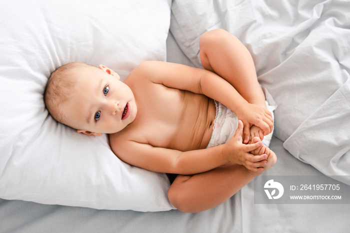 Top view of little baby in diaper laying on back and grabbing feet with hands and looking at camera