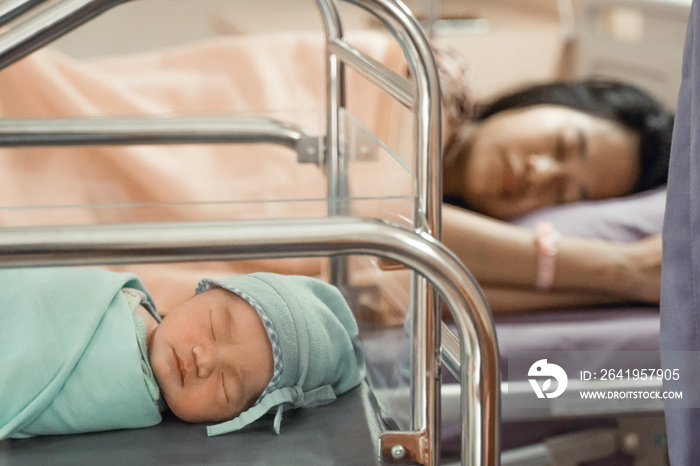 Newborn baby laying in crib with his mother lying on bed side in the hospital