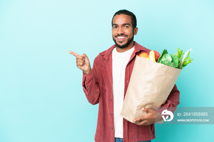 Young latin man holding a grocery shopping bag isolated on blue background pointing finger to the si