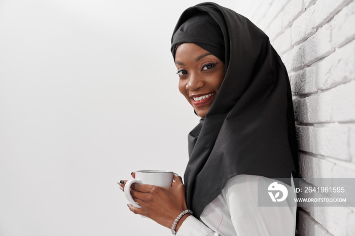 Muslim woman in hijab leaning at wall, holding white cup.