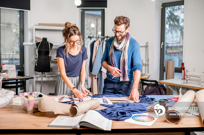 Couple of fashion designers working with fabric and clothing sketches at the studio full of tailorin