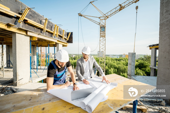 Engineer with worker in uniform working with architectural drawings and laptop at the table on the c