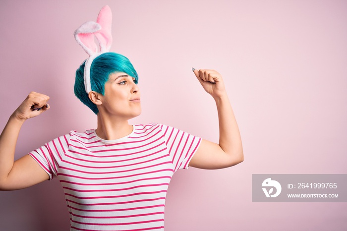 年轻女性，时尚蓝色头发，在粉色背景下戴着复活节兔子耳朵，手臂肌肉发达