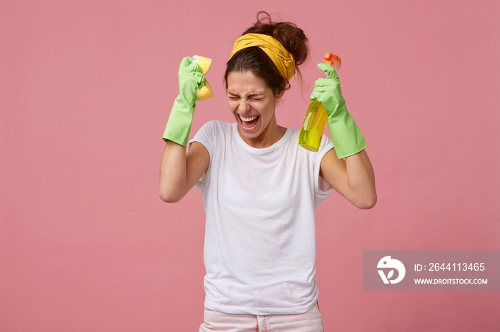Dissatisfied young woman holding sponge and detergent wearing gloves, yellow headband on head and wh