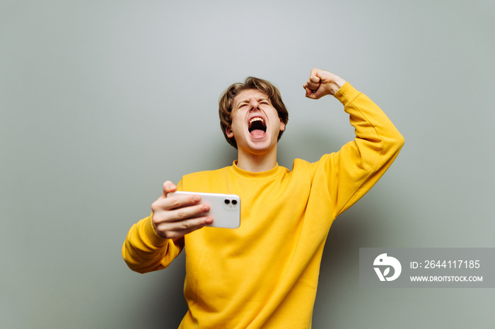 Emotional young man in yellow clothes rejoices in victory with a smartphone in his hands on a gray b