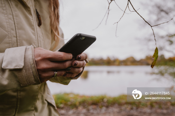 Close-up of phone in woman`s hands. River at cloudy autumn day in the background.