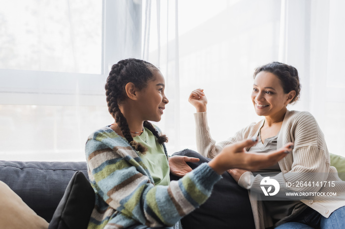 teenage african american girl pointing with hand while talking to smiling mother on sofa