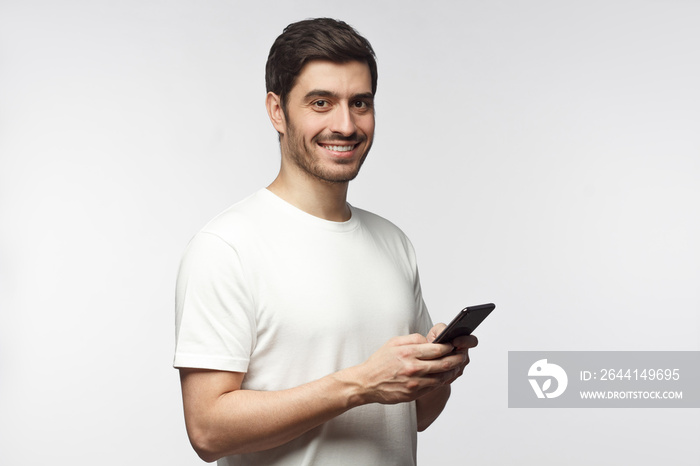 Young man looking and smiling at the camera with cheerful expression while texting to friend, using 