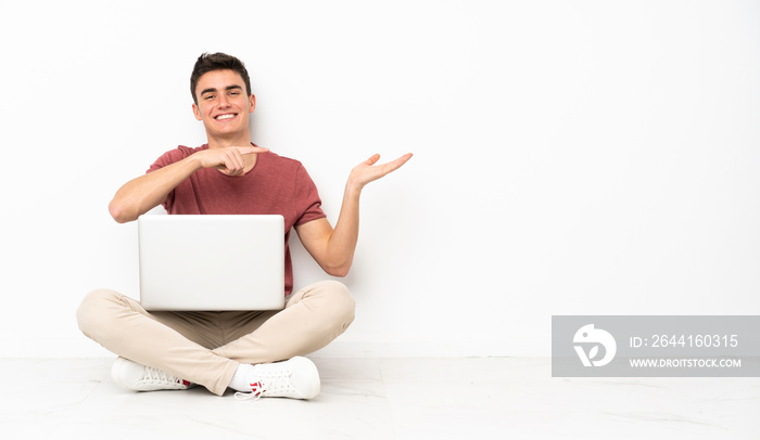 Teenager man sitting on the flor with his laptop holding copyspace imaginary on the palm to insert a