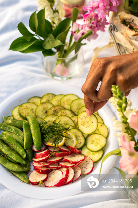 Black woman holding a cucumber slice from crudites platter while enjoying a luxury sunset picnic in 