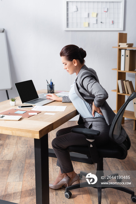 Tired businesswoman with pain in back holding hand on hip, while sitting on office chair in office