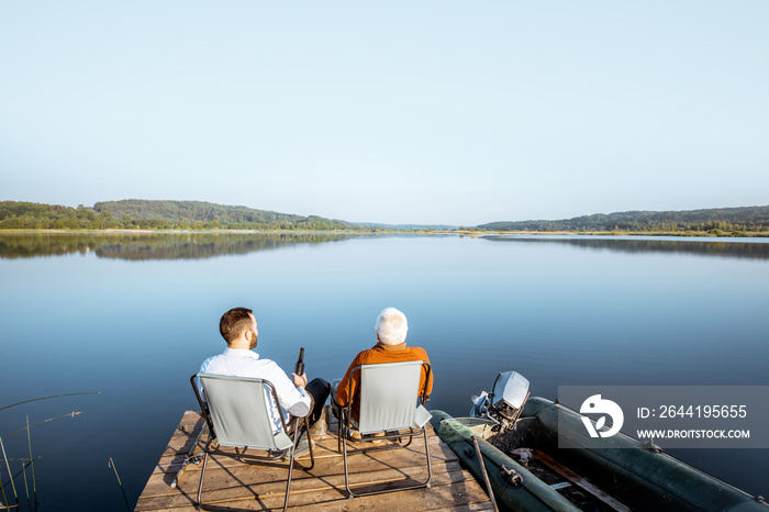 Grandfather with adult son enjoying beer, sitting together on the pier while fishing on the lake ear