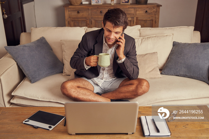 Young man teleworking from home in video conference, sitting on the couch in a suit and shorts. Drin