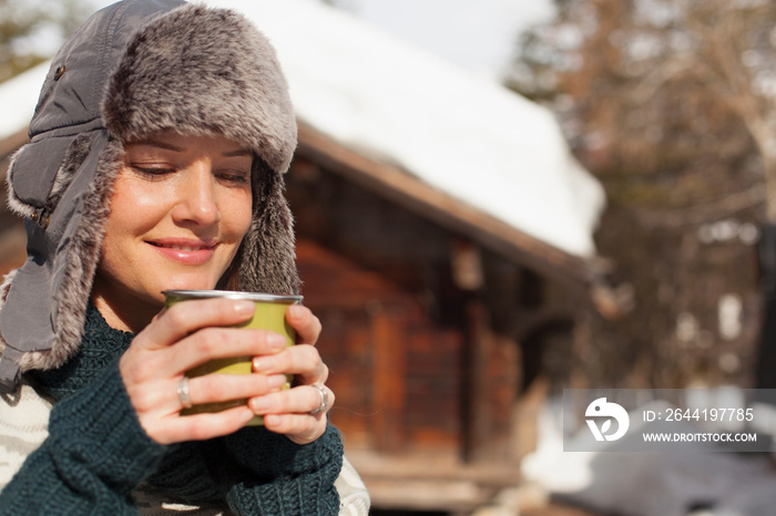 Smiling woman in fur hat drinking coffee outside cabin