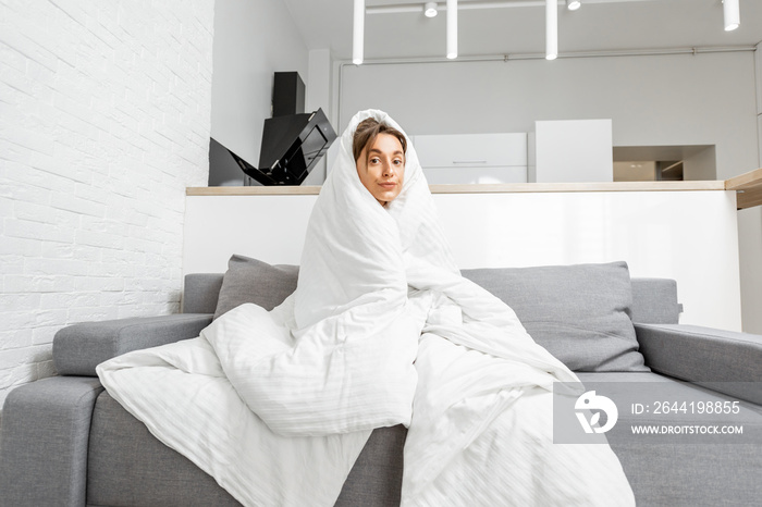 Sleepy woman covered with a white blanket on the couch at home, wide interior view. Concept of self-