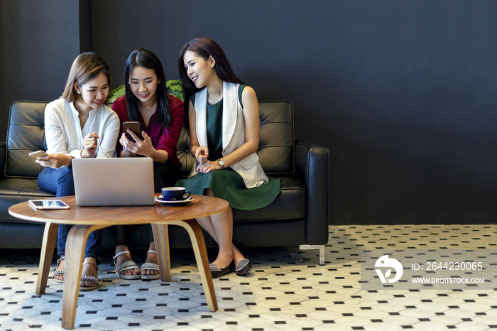 Group of charming beautiful Asian women using smartphone and laptop, chatting on sofa at cafe, moder