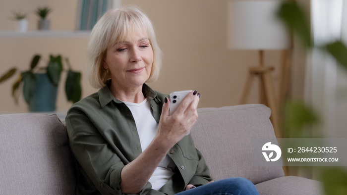 Old Caucasian woman 60s elderly mature lady sits at home on sofa with mobile phone reads news receiv