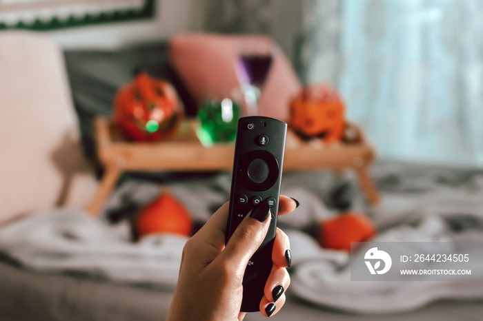 Woman hand with black manicure holding tv remote with Halloween decor in background, Halloween movie