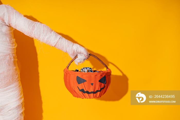 Child in mummy costume holds basket in the form of pumpkin with sweets in his hands Halloween trick 