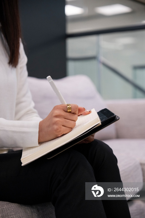 Midsection of woman sitting on sofa and writing in notepad