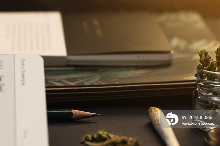 Jar of cannabis buds, gold lid, notebooks, joint on black background