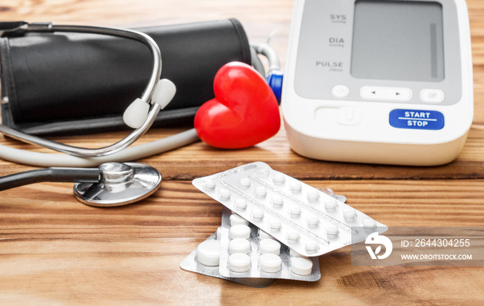 Pills with tonometr, stethoscope and red heart on wooden table.