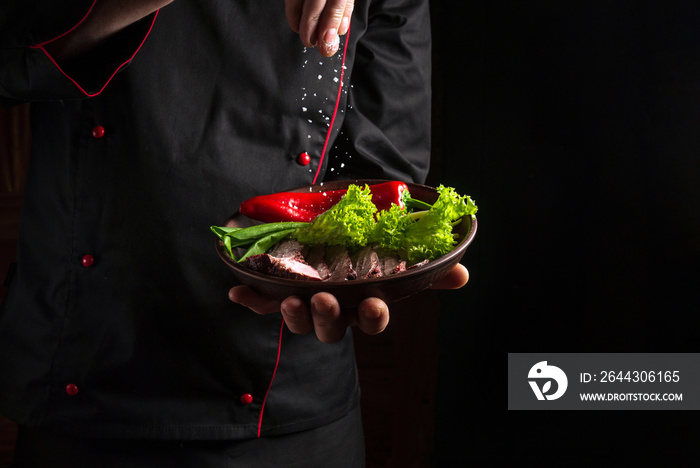 Chief sprinkles salt on a sliced steak with beef and vegetables in a plate. The concept of serving dishes to order