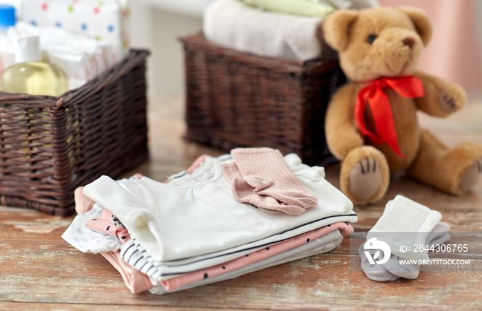 babyhood and clothing concept - baby clothes, teddy bear toy and baskets on wooden table at home