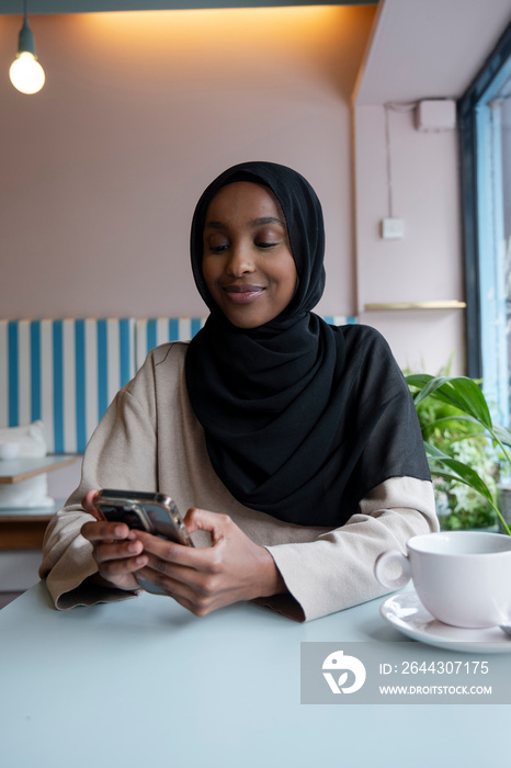 Smiling young woman in hijab using phone in cafe