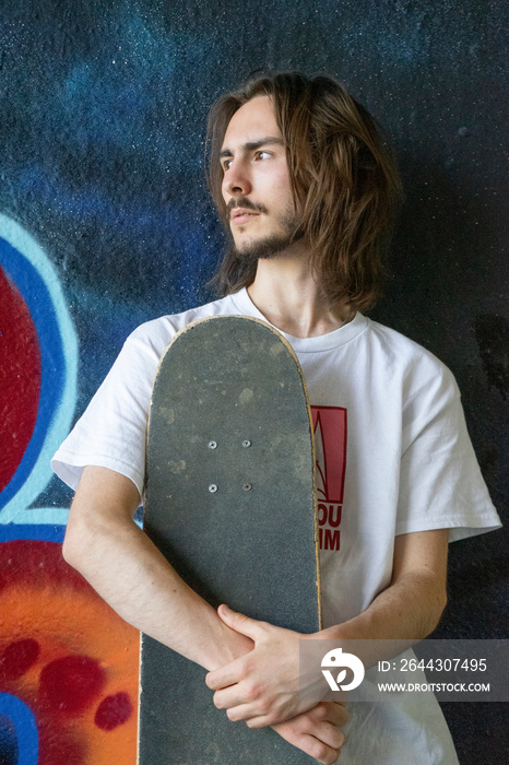 Young man standing against graffiti wall and holding skateboard