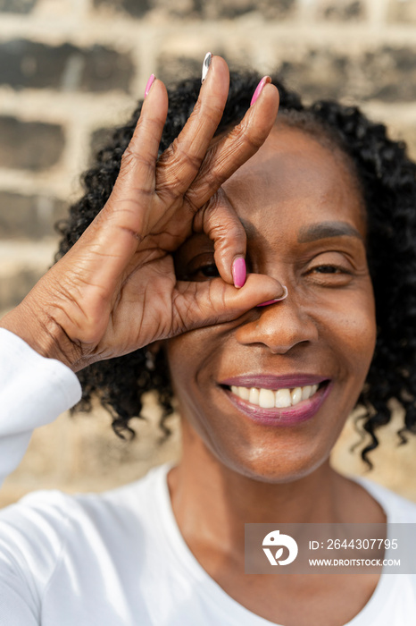 Portrait of woman looking through OK sign made with fingers against brick wall