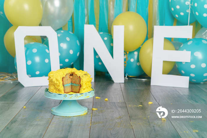 Festive background decoration for birthday celebration with yellow cake, letters saying one and blue balloons in studio. Cake smash first year concept.