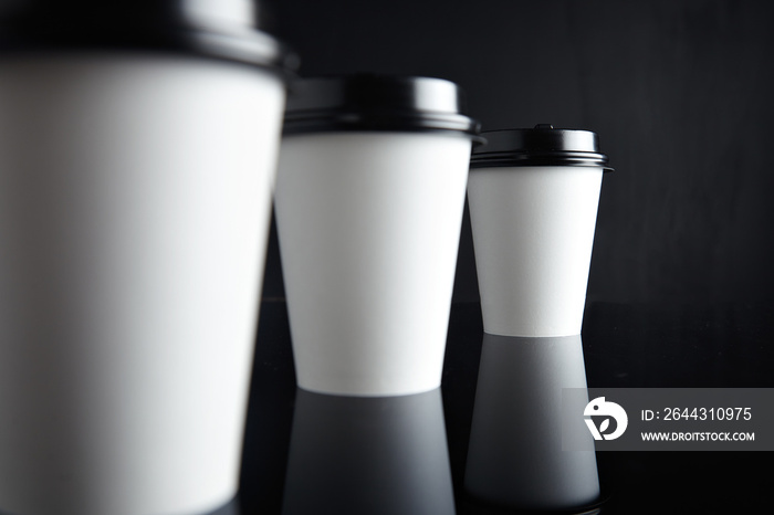 One focused paper cup for hot drinks behind two unfocused presented on black back and mirrored, closed with caps