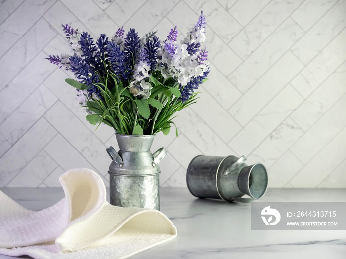 Rustic metal milk can filled with artificial purple lavender and lilacs for a simple spring country home decor with still life photography and a marble counter with a herringbone tile background.