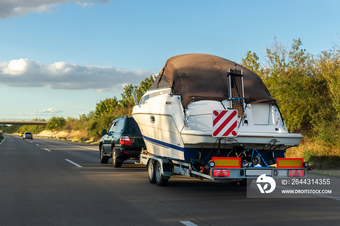 Big luxury cabin motorboat cruiser yacht trailer SUV carrier go to river lake on highway road sunset summer light. Luxury rich fishing leisure recreation lifestyle. Service maintenance transportation