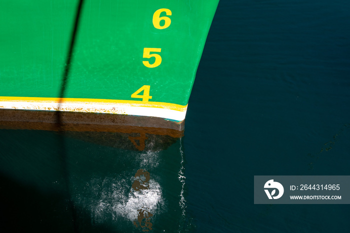 The bow of a large green ship with brown and white stripes at the waterline. The numbers 4, 5, and 6 are painted on the boat in yellow paint as a measurement of meters of water depth or load line.