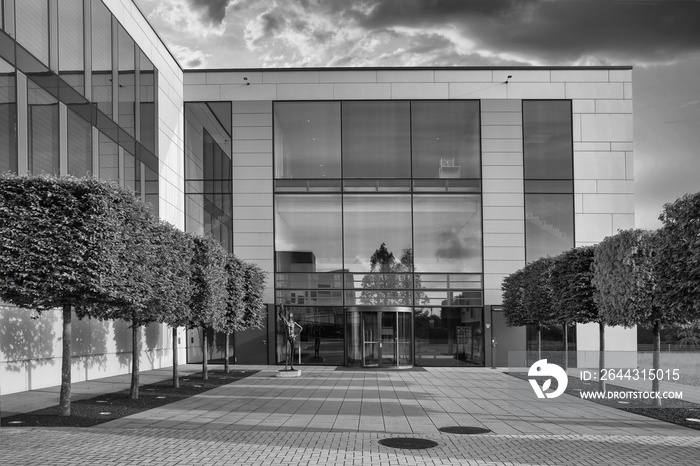 Greyscale shot of a modern building with glass windows and trees on the front