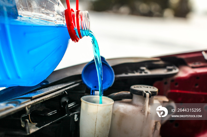 Detail on pouring antifreeze liquid screen wash into dirty car from blue and red anti freeze water container.