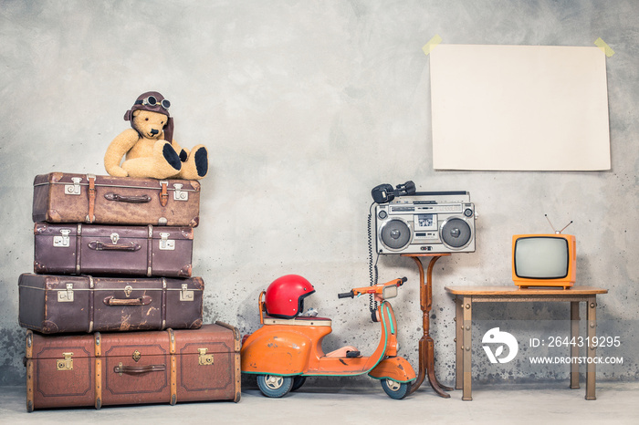 Retro Teddy Bear toy in aviator’s hat, aged travel valises, children’s pedal scooter, helmet, cassette boombox with headphones, TV, paper poster blank on concrete wall. Vintage style filtered photo