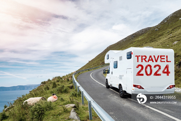 White motorhome with sign travel 2024 moving on a small mountain road and beautiful country side with ocean in foreground. Low cloudy sky. Travel with camper concept. Trip to nature.