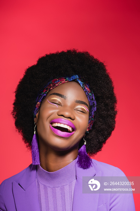 laughing african american woman in purple stylish outfit isolated on red