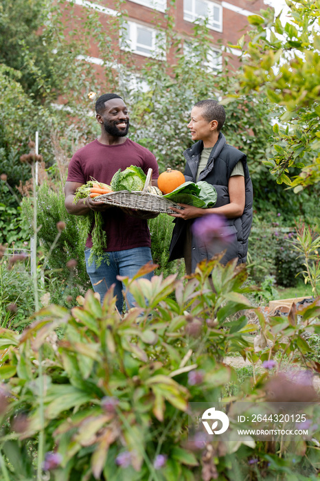 Portrait of smiling couple holding basket with fresh vegetables in urban garden