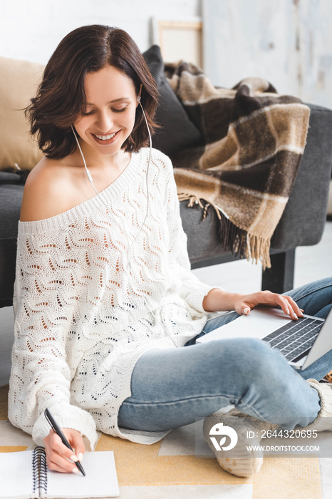 cheerful girl with earphones studying online with laptop and notebook at home