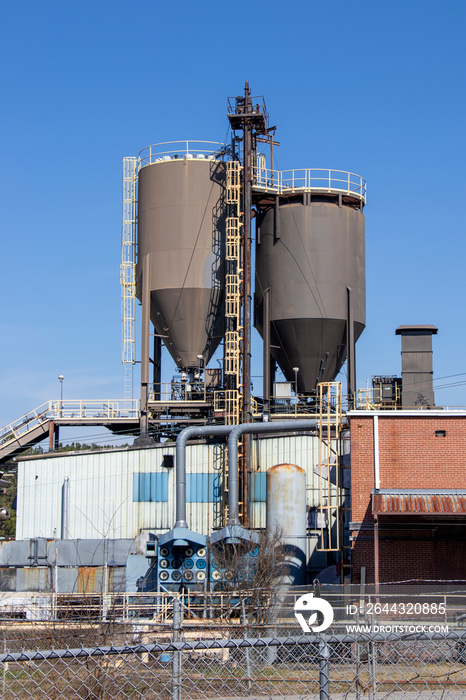 Vertical exterior of a brick and metal industrial building with storage silos, a dust collection system, and a conveyor behind a barbed wire fence.